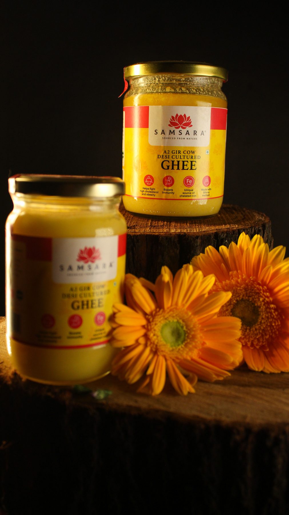 Two Samsara ghee jars on stacked wood with orange daisies